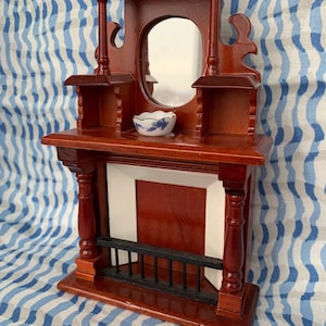 May include: Miniature wooden fireplace mantel with a mirror, shelves, and a small decorative bowl. The mantel is stained a dark reddish-brown color with a white and black fireplace insert. A small gold decorative piece sits on top.