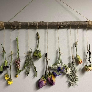 May include: A rustic wooden branch with dried flowers and herbs hanging from it. The flowers are in shades of purple, pink, yellow, and white. The herbs are green and brown.
