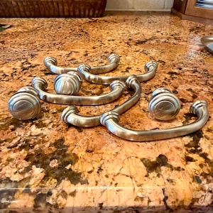 May include: A collection of brushed nickel cabinet pulls and knobs. The pulls have a curved handle design with decorative detailing, and the knobs feature a round, fluted shape. These hardware pieces are designed for kitchen or bathroom cabinetry.