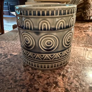 May include: A cylindrical, gray ceramic planter with a repeating pattern of arches, concentric circles, and geometric shapes. The planter has a textured surface and a slightly flared rim. The design is in a darker shade of gray.