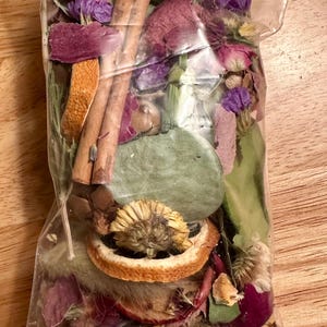 以下が含まれることがあります： 紫色の花、緑の葉、オレンジのスライス、シナモンスティックなど、乾燥した植物が詰まった透明なビニール袋です。