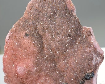 May include: A natural mineral specimen featuring a blend of pink and brown hues. The rock has a rough, textured surface with small, sparkling crystal formations. The overall color is a soft pink, with areas of darker brown and black.