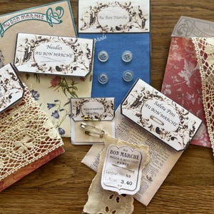 Op de afbeelding: Assortiment van vintage naaibenodigdheden, waaronder kant, naalden, veiligheidsspelden en knopen. De items zijn verpakt in decoratieve dozen en kaarten met Franse tekst en illustraties. Het kant is crèmekleurig en de knopen zijn transparant.