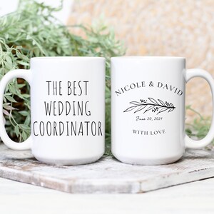 Może przedstawiać: Dwa białe ceramiczne kubki z czarnym tekstem. Jeden kubek ma napis "The Best Wedding Coordinator", a drugi kubek ma napis "Nicole & David" z kwiatowym wzorem i datą "June 20, 2021" oraz słowami "With Love".