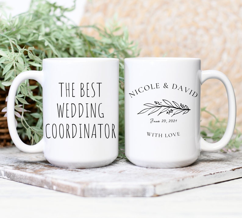 Może przedstawiać: Dwa białe ceramiczne kubki z czarnym tekstem. Jeden kubek ma napis "The Best Wedding Coordinator", a drugi kubek ma napis "Nicole & David" z kwiatowym wzorem i datą "June 20, 2021" oraz słowami "With Love".