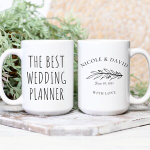 May include: Two white ceramic mugs with black text. One mug says "The Best Wedding Planner" and the other mug says "Nicole & David" with a floral design and the date "June 20, 2021" and the words "With Love".