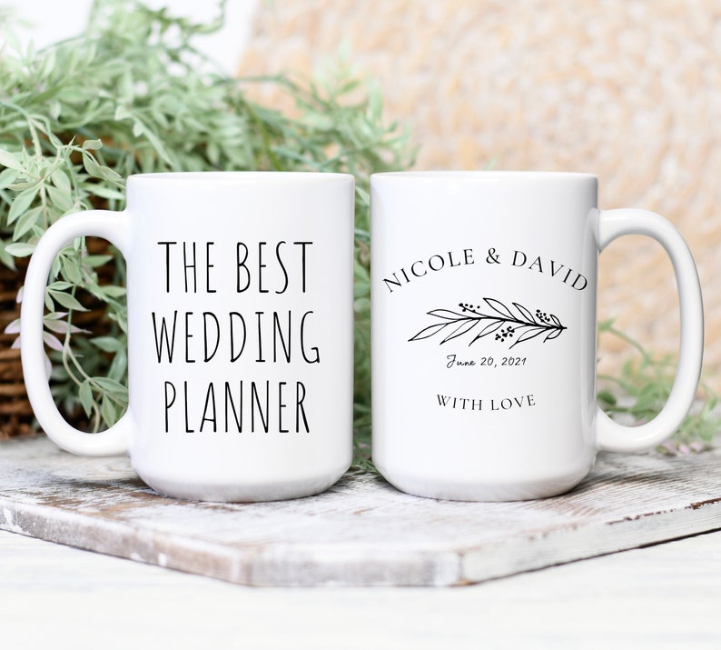 May include: Two white ceramic mugs with black text. One mug says "The Best Wedding Planner" and the other mug says "Nicole & David" with a floral design and the date "June 20, 2021" and the words "With Love".
