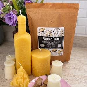 May include: A brown paper bag of Pioneer Blend coffee labelled 400g, surrounded by yellow beeswax candles in bottle and pillar shapes, small cream-coloured candles, and a pink plate with brown coffee cubes and a silver spoon.