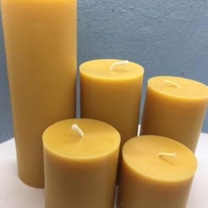 May include: A collection of five cylindrical beeswax candles in varying heights, each featuring a smooth, golden-yellow surface and a single white cotton wick centred at the top. The candles are arranged in a cluster with the tallest positioned at the back.