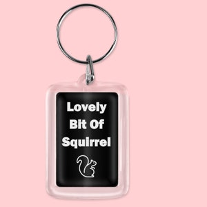 May include: A clear acrylic keychain with a black background and white text that reads "Lovely Bit Of Squirrel". A white silhouette of a squirrel is below the text.