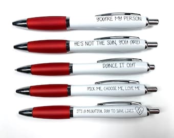May include: Five white pens with red caps and black ink. Each pen has a different quote printed on it: "You're my person", "He's not the sun, you are!", "Dance it out", "Pick me, choose me, love me", and "It's a beautiful day to save lives".