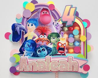 May include: A colorful cake topper featuring characters from the movie "Inside Out", with the number 4 and the name "Analeah". The design includes a rainbow, colorful circles, and a candy-filled display.