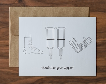 May include: A white card with black line drawings of a medical boot, crutches, and a bandaged arm. The text "thanks for your support" is below the drawings. The card is inside a brown envelope.