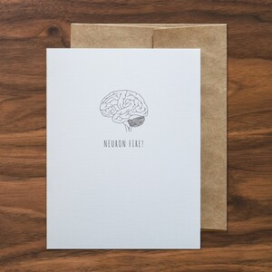 May include: A white greeting card with a black line drawing of a brain and the words "NEURON FIRE!". The card is paired with a brown envelope. The card and envelope are on a wooden surface.