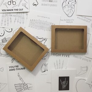 Puede incluir: Dos cajas de cart&oacute;n marr&oacute;n con ventanas transparentes se muestran entre varias tarjetas de felicitaci&oacute;n en blanco y negro. Las tarjetas presentan ilustraciones anat&oacute;micas y frases basadas en juegos de palabras como "YOU MADE THE CUT" y "HIGH FIVE".