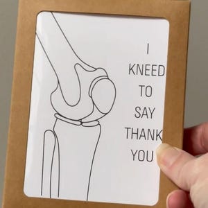 Puede incluir: Una tarjeta marr&oacute;n con una inserci&oacute;n blanca que presenta un dibujo lineal negro de una articulaci&oacute;n de la rodilla. El texto "I KNEED TO SAY THANK YOU" est&aacute; impreso en negro. La tarjeta est&aacute; sujeta por una mano.
