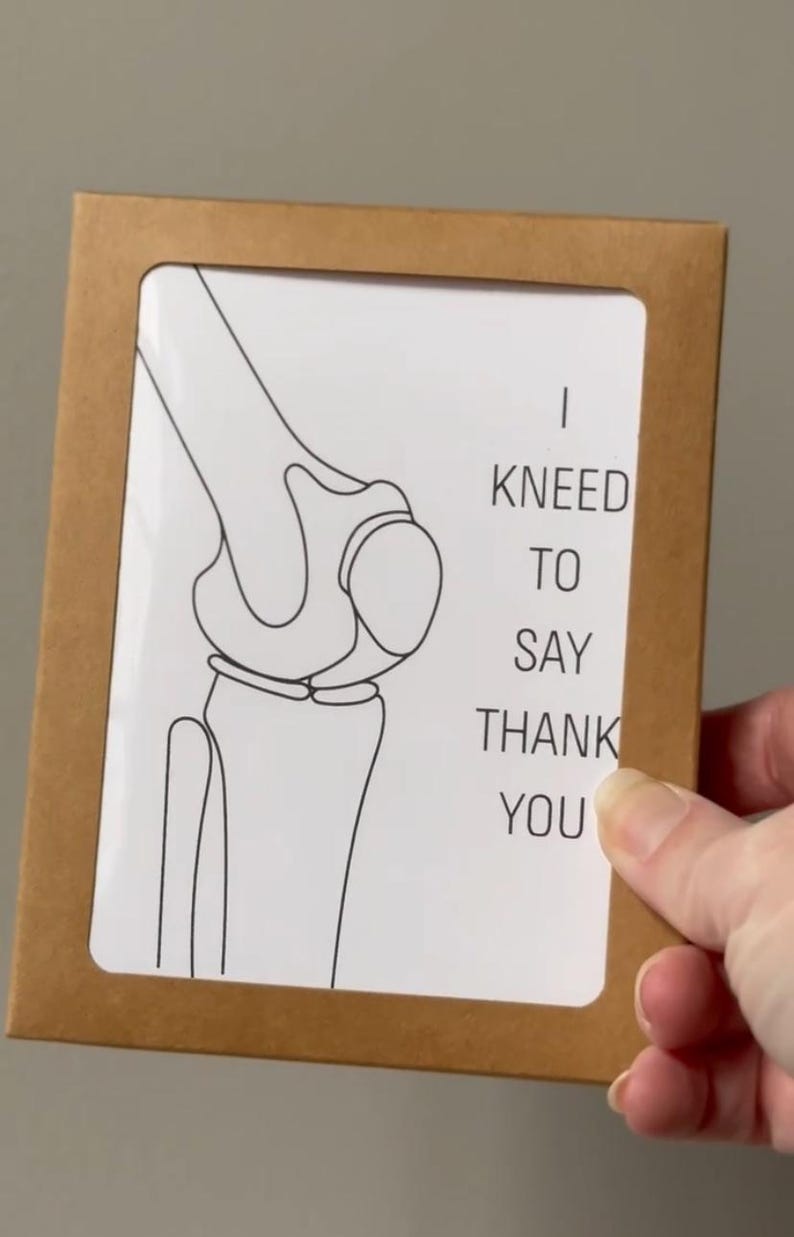 Puede incluir: Una tarjeta marr&oacute;n con una inserci&oacute;n blanca que presenta un dibujo lineal negro de una articulaci&oacute;n de la rodilla. El texto "I KNEED TO SAY THANK YOU" est&aacute; impreso en negro. La tarjeta est&aacute; sujeta por una mano.
