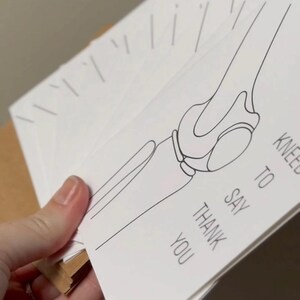 Puede incluir: Una pila de tarjetas de agradecimiento blancas con un dibujo lineal negro de una articulaci&oacute;n de la rodilla. El texto "KNEE TO SAY THANK YOU" est&aacute; impreso en la tarjeta. Las tarjetas se sostienen en una mano.