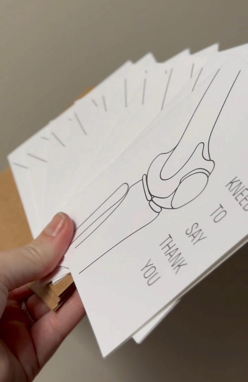 Puede incluir: Una pila de tarjetas de agradecimiento blancas con un dibujo lineal negro de una articulaci&oacute;n de la rodilla. El texto "KNEE TO SAY THANK YOU" est&aacute; impreso en la tarjeta. Las tarjetas se sostienen en una mano.
