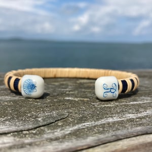 Puede incluir: Un brazalete tejido con extremos de cerámica azul y blanca. Un extremo tiene un diseño de concha marina, y el otro extremo tiene un monograma de la letra "R" en azul.
