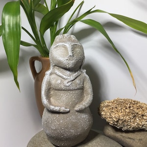 May include: A gray stone sculpture of a woman with a simple, stylized design. The sculpture is standing on a rock and is surrounded by green plants.