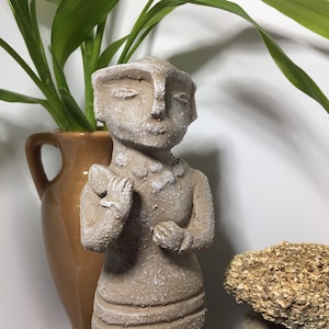 May include: A small, gray, ceramic statue of a person with a stylized face and a simple, pleated dress. The statue is standing on a rock.