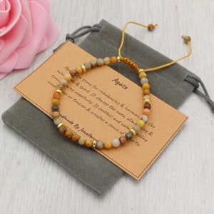 Könnte beinhalten: Agat-Armband mit braunen, gelben und weißen Perlen. Das Armband liegt auf einer braunen Karte mit dem Text "Agate is a stone for rebalancing & harmonizing the mind, body & spirit. It cleanses the aura, eliminating negativity. It soothes and calms healing inner anger or tension & creates a sense of clarity." und "Handmade By Jewellerud".