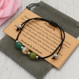 Könnte beinhalten: Ein schwarzes Kordelarmband mit drei Edelsteinperlen in Grün, Weiß und Gelb, akzentuiert mit silberfarbenen Perlen. Das Armband wird auf einem grauen Samtbeutel mit einer Karte mit der Aufschrift "Prosperity" präsentiert.