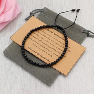 May include: Black tourmaline beaded bracelet with a drawstring closure, presented on a grey pouch with a tag. The tag reads "Black Tourmaline" and describes its properties. Handmade by Jewelleryden.
