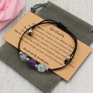 May include: A black cord bracelet featuring clear, purple, and white gemstone beads. The bracelet is presented on a small brown card with text about spirituality and a grey pouch. The card reads "Handmade By Jewelleryden."