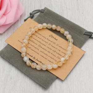 Könnte beinhalten: Ein Citrin-Edelsteinarmband in hellem Gelb, das auf einem grauen Samtbeutel und einer beigen Karte liegt. Die Karte enthält Text, der die Eigenschaften von Citrin beschreibt. Handgefertigt.