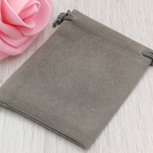 May include: A small, grey velvet drawstring pouch with a matching cord closure. The pouch is rectangular and has a soft, textured appearance. A pink rose is partially visible in the background, adding a touch of colour.