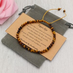 May include: A Tigers Eye beaded bracelet with a tan cord, presented on a grey pouch with a small tag. The tag details the stone's properties. The bracelet is handmade.