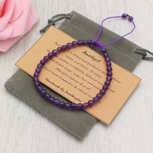 May include: A handmade amethyst bead bracelet with a purple cord, displayed on a grey velvet pouch. A small card with the word "Amethyst" and product information is visible. The text "Handmade By Jewelleryden" is also visible.