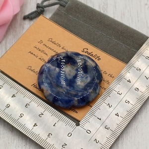 May include: A blue and white sodalite stone, flower-shaped, rests on a wooden card with text. A ruler shows measurements in centimetres. The stone is next to a gray drawstring bag. The card reads "Sodalite".