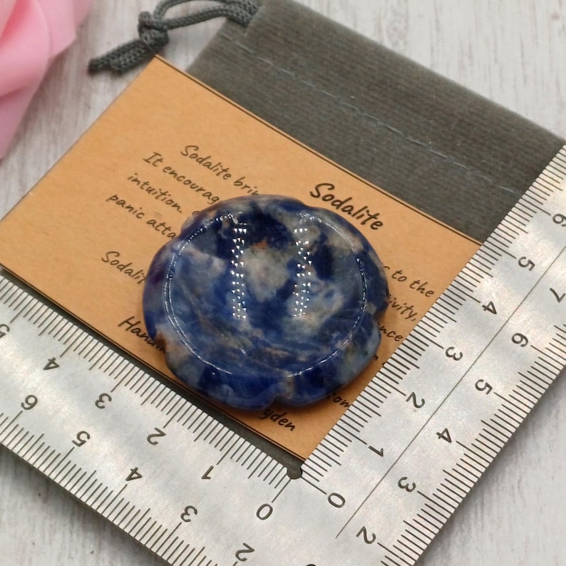 May include: A blue and white sodalite stone, flower-shaped, rests on a wooden card with text. A ruler shows measurements in centimetres. The stone is next to a gray drawstring bag. The card reads "Sodalite".