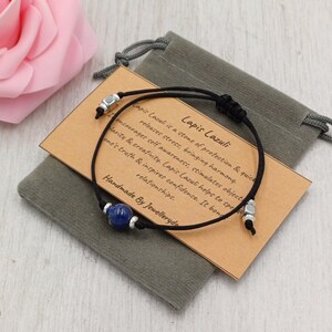 Peut inclure: Un bracelet en cordon noir avec une perle de lapis-lazuli bleue et des détails argentés. Le bracelet est présenté sur une petite carte marron avec du texte et une pochette en velours grise. La carte indique "Lapis Lazuli".