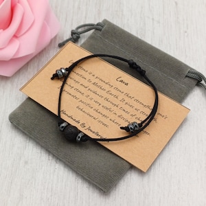 May include: A black lava stone bracelet with silver beads and a black cord. The bracelet rests on a grey velvet pouch with a tag that reads "Lava". The tag also has text describing the stone's properties.