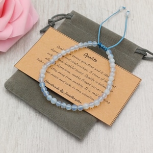 Könnte beinhalten: Ein zartes Opalit-Edelsteinarmband mit hellblauen Perlen, präsentiert auf einem grauen Beutel mit einem Etikett. Das Etikett trägt die Aufschrift "Opalit" und beschreibt die Eigenschaften des Edelsteins. Handgefertigt von Jewelleryden.