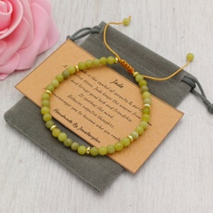 Könnte beinhalten: Ein grünes Jade-Armband mit goldenen Perlen und einer braunen Karte mit der Aufschrift "Jade" und einer Beschreibung der Eigenschaften des Steins. Das Armband befindet sich in einem grauen Samtbeutel.