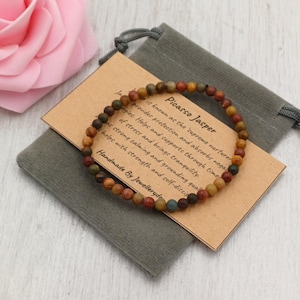 Puede incluir: Una pulsera de cuentas de Jaspe Picasso descansa sobre una bolsa de terciopelo gris, acompañada de una etiqueta con texto. La pulsera presenta pequeñas cuentas redondas en tonos marrón, verde y rojo. Una rosa rosa está en el fondo.