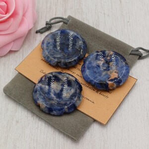 May include: Three blue sodalite stones with a scalloped edge, on a grey pouch with a drawstring. A small tan card with text is visible. A pink rose is in the background.