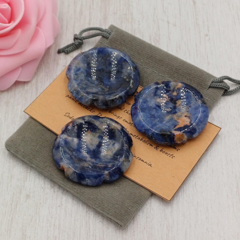 May include: Three blue sodalite stones with a scalloped edge, on a grey pouch with a drawstring. A small tan card with text is visible. A pink rose is in the background.