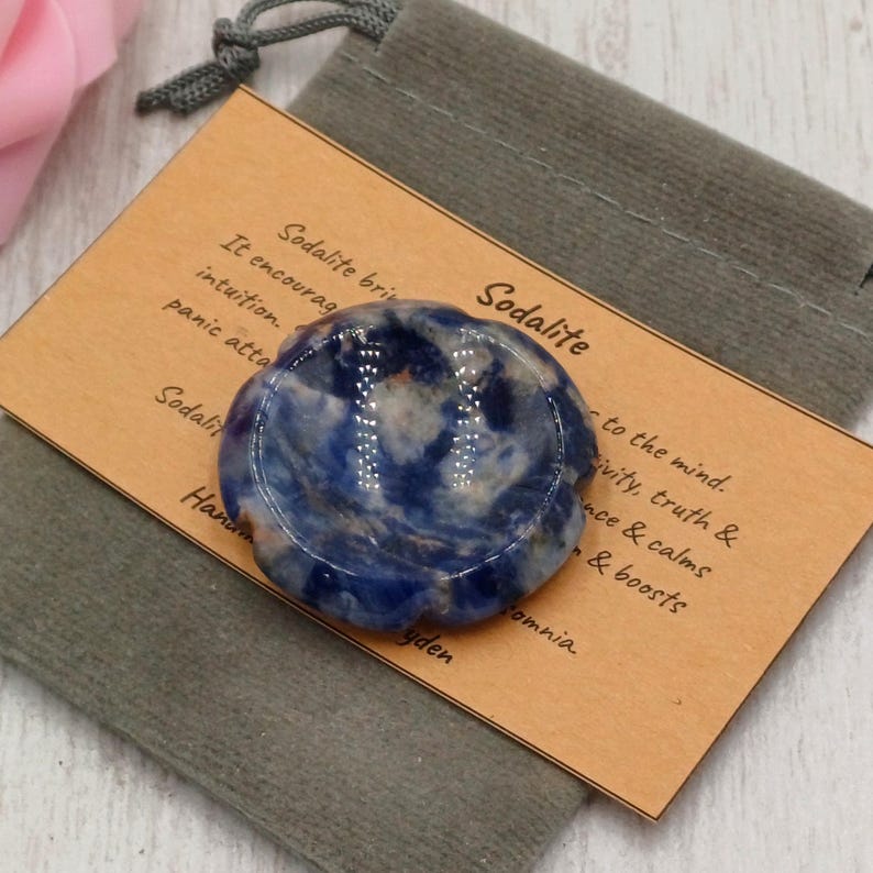 May include: A blue sodalite stone with white veining, presented on a tan card with text. The stone is in a grey pouch. The card reads "Sodalite" and describes its properties.