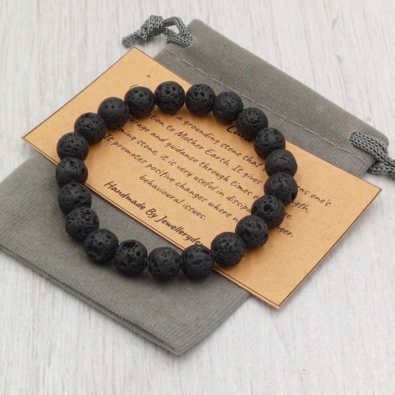 Puede incluir: Pulsera de cuentas de roca volc&aacute;nica negra con una tarjeta marr&oacute;n que dice "Una piedra de conexi&oacute;n a tierra que da conexi&oacute;n a la Madre Tierra. Da coraje y gu&iacute;a en tiempos de cambio. Promueve cambios positivos donde sea necesario. Hecho a mano por Jewelleryden."
