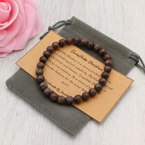 May include: Brown Snowflake Obsidian bracelet with a brown card that reads "Snowflake Obsidian is a strong protection stone, acting as a shield against negativity. Obsidian is known for its ability to help with mental stress and tension. As a stone of protection, it is calming and soothing. It provides balance for the mind, body and spirit. Handmade by Jewell."