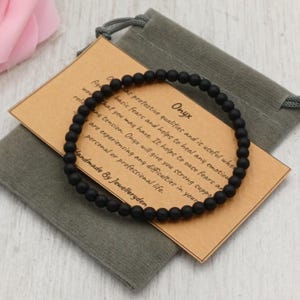 May include: A black onyx beaded bracelet, presented with a small grey drawstring pouch and a tan card. The card features the word "Onyx" and text detailing the stone's properties. The bracelet is handmade.
