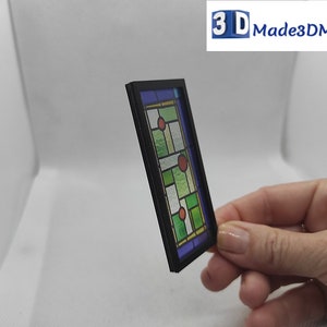 May include: A miniature stained glass window with a black frame. The glass features a geometric design with green, blue, and red accents.