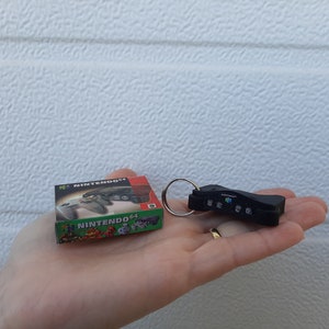Puede incluir: Un llavero negro de Nintendo 64 con un anillo de llave plateado. El llavero tiene la forma de una consola Nintendo 64. El llavero está al lado de una pequeña caja con el logotipo de Nintendo 64 y las palabras "N64 Nintendo 64" en ella.