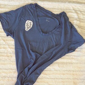 Puede incluir: Una camiseta azul con un dise&ntilde;o floral blanco en el lado izquierdo del pecho. El dise&ntilde;o es un simple dibujo de l&iacute;nea de una rama con hojas y flores.